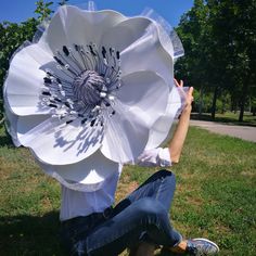 a woman sitting in the grass holding up a large white flower with black and silver details