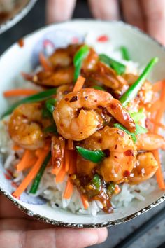 a person holding a bowl with shrimp and carrots on rice in it, while another hand is holding the bowl