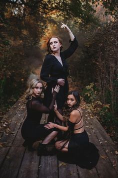 three women in black dresses posing on a wooden platform with trees and leaves behind them