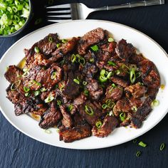 a white plate topped with meat and green onions next to a bowl of chopped scallions