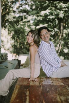 a man and woman sitting on top of a wooden table in front of some trees