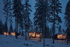 the cabin is lit up at night in the snow covered forest with lights shining on it