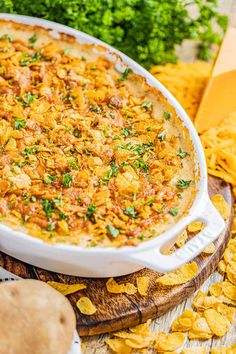 a casserole dish with cheese and herbs in it on a cutting board next to chips