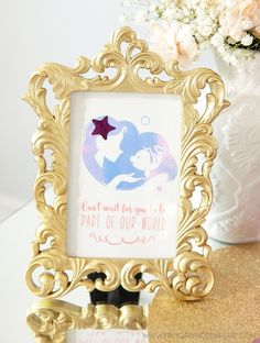 there is a gold frame with a little mermaid in it on top of a table