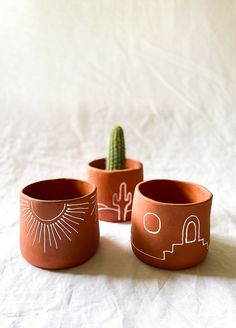 three clay pots with designs on them and a cactus in the middle, sitting on a white sheet