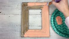 a person is painting the frame on a white wooden table with a green and pink plate