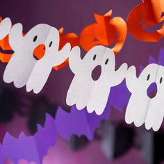 paper ghost garlands hanging on a purple and orange background with halloween decorations in the foreground