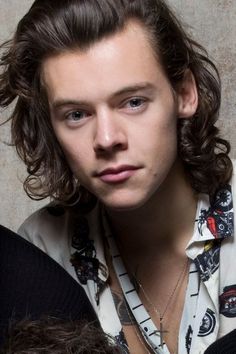 a young man with long hair wearing a white shirt and looking at the camera while standing in front of a wall