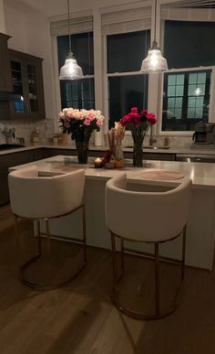 two white stools sitting in front of a kitchen island with flowers on it and lights hanging from the ceiling