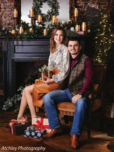 a man and woman sitting on a chair in front of a fireplace with christmas decorations