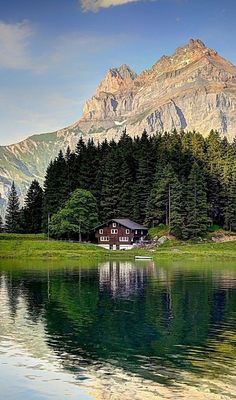 a house on the shore of a lake in front of a mountain with pine trees