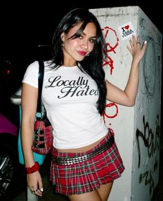 a woman in a white shirt and red plaid skirt holding up a cell phone while standing next to a wall with graffiti on it