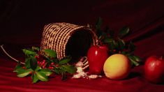 an apple, orange and pomegranate on a red cloth with holly leaves