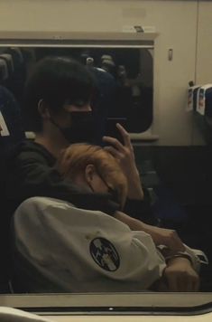 a man and woman sitting on a train looking at their cell phones while wearing masks