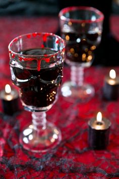 two glasses filled with liquid sitting on top of a table next to candles and a red cloth