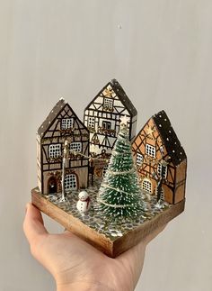 a hand holding a miniature christmas village with snow on the ground and trees in front