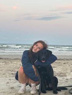 a woman kneeling down next to a black dog on a beach with the ocean in the background