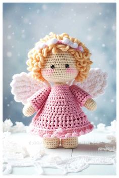 a crocheted doll with blonde hair wearing a pink dress and angel wings on top of snow covered ground