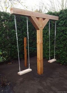 a wooden swing set with two swings attached to it's sides and some bushes in the background