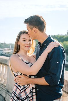 a man and woman standing next to each other in front of a bridge with the water behind them