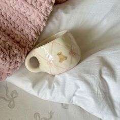 a white cup sitting on top of a bed next to a pink blanket and pillow