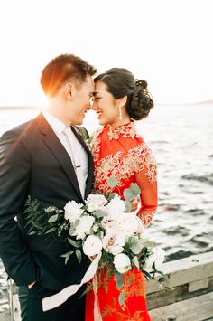 a man and woman standing next to each other near the ocean with flowers in front of them