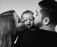 a black and white photo of a man kissing a woman's face while holding a baby