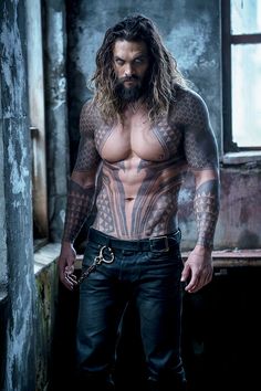 a man with long hair and tattoos on his chest standing in an old building, wearing black jeans