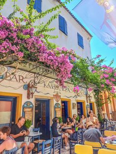 people sitting at tables in front of a building with purple flowers on the outside wall