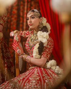 a woman in a red and gold bridal outfit sits on a chair with her hands clasped