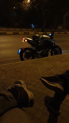 a person sitting on the ground with their feet up next to a parked motorcycle at night