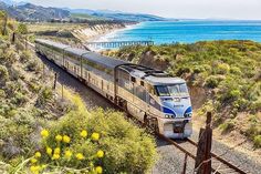 a train traveling down tracks next to the ocean and beach with mountains in the background