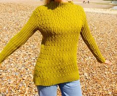 a woman is standing on the beach wearing a green sweater and jeans with her arms outstretched