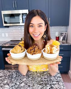 a woman holding three cupcakes on a tray in front of her face and smiling at the camera