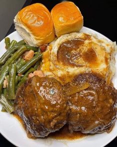 a white plate topped with meat covered in gravy next to green beans and rolls