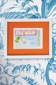 an orange and white frame with a painting on it that says demmar in front of a blue floral wallpaper