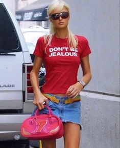 a woman in red shirt and denim shorts carrying a pink handbag while standing next to a white truck