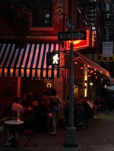 people are sitting at tables on the sidewalk in front of a restaurant with neon signs