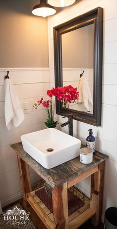 a white sink sitting under a bathroom mirror next to a wooden stand with flowers on it