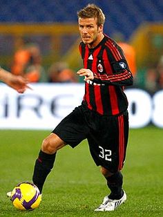 a man in black and red uniform kicking a soccer ball on the field with other people watching