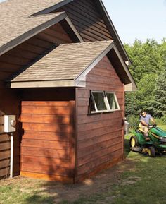 a man on a lawn mower in front of a small wooden building with shingled roof