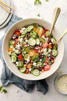 a white bowl filled with cucumber, tomato and feta cheese salad next to two silver spoons