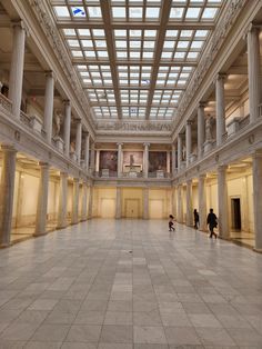 two people are walking in an empty building with columns and glass ceiling above the floor