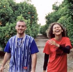 two people standing next to each other on a dirt road near trees and oranges