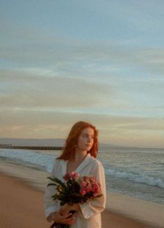 a woman standing on top of a beach next to the ocean holding a bouquet of flowers