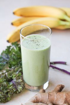 a glass filled with green smoothie next to some bananas and kale on the table