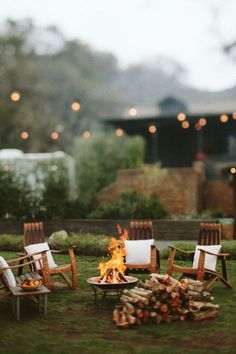 a fire pit with chairs around it in the middle of some grass and lights hanging overhead