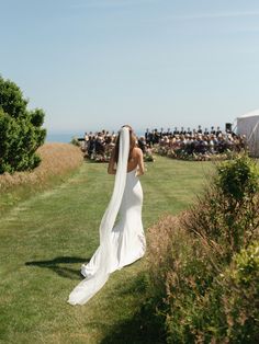 a woman in a wedding dress is walking down the grass with her veil blowing in the wind