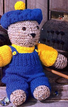 a crocheted teddy bear wearing a blue hat and overalls sitting on a bench