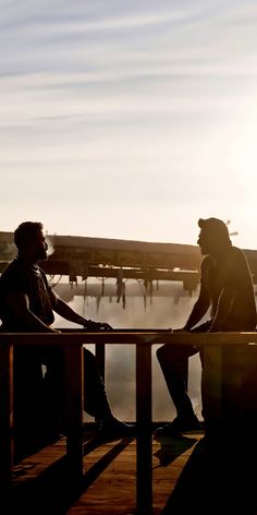 two people are sitting on a dock near the water and one is talking to another person